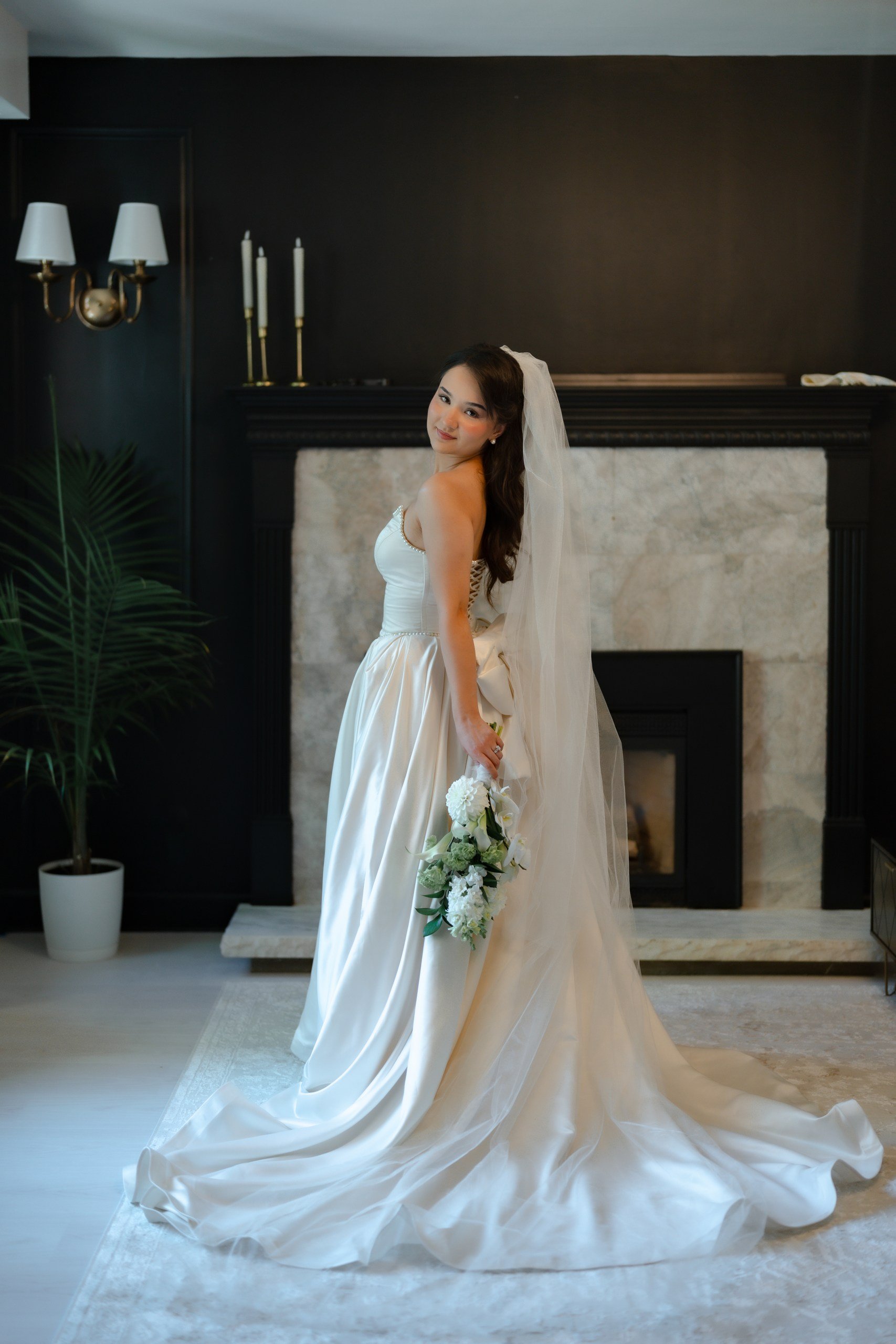 A bride in a strapless white wedding gown with a long train, holding a bouquet of white flowers, stands indoors in front of a black fireplace with a marble mantel. She has long dark hair with soft waves and wears a veil, looking over her shoulder and