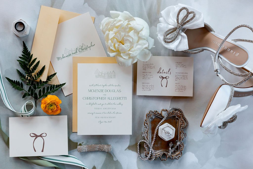 Wedding invitation suite with details card, floral decorations, pearl jewelry, white and silver high-heeled shoes with bows, white flowers, and greenery on a light surface.