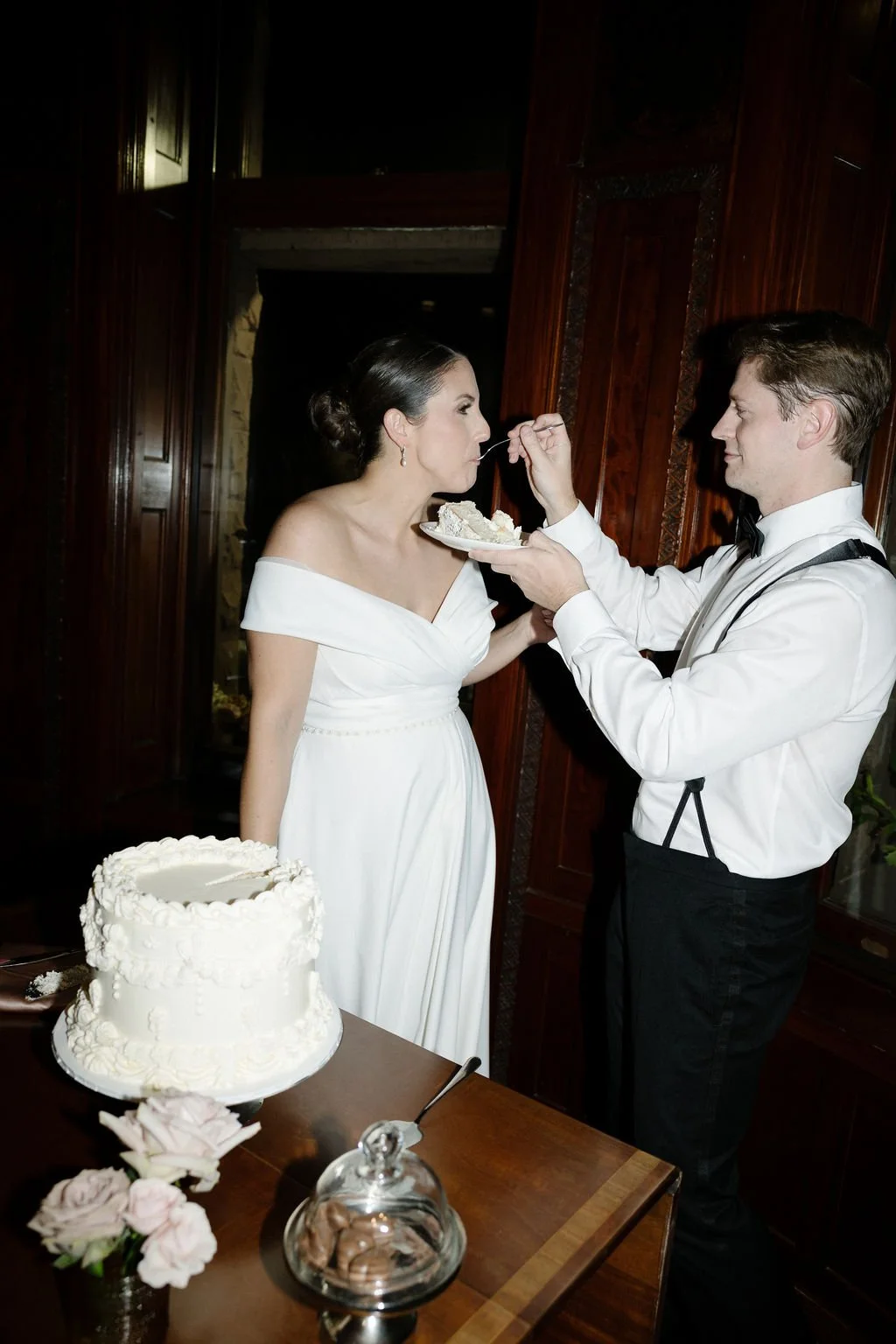 A bride in a white wedding dress being fed cake by a groom in black tuxedo pants, white shirt, and suspenders at a wedding reception, with a wedding cake, flowers, and a jar of chocolates on the table in front of them.