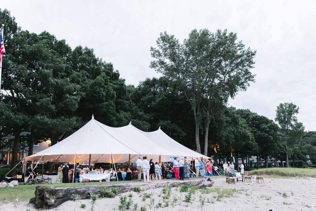 Outdoor gathering under a large white tent with people, tables, and chairs on a sandy beach, surrounded by green trees.
