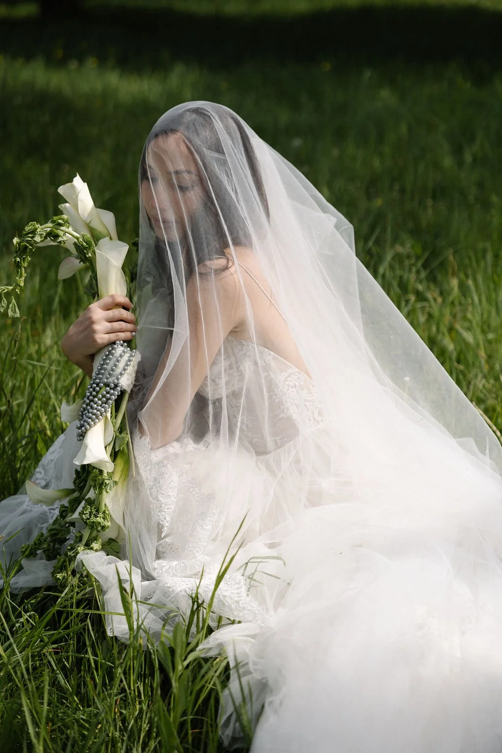 A bride sitting in a grassy field, wearing a white wedding dress and veil, holding a bouquet of calla lilies and pearl jewelry.