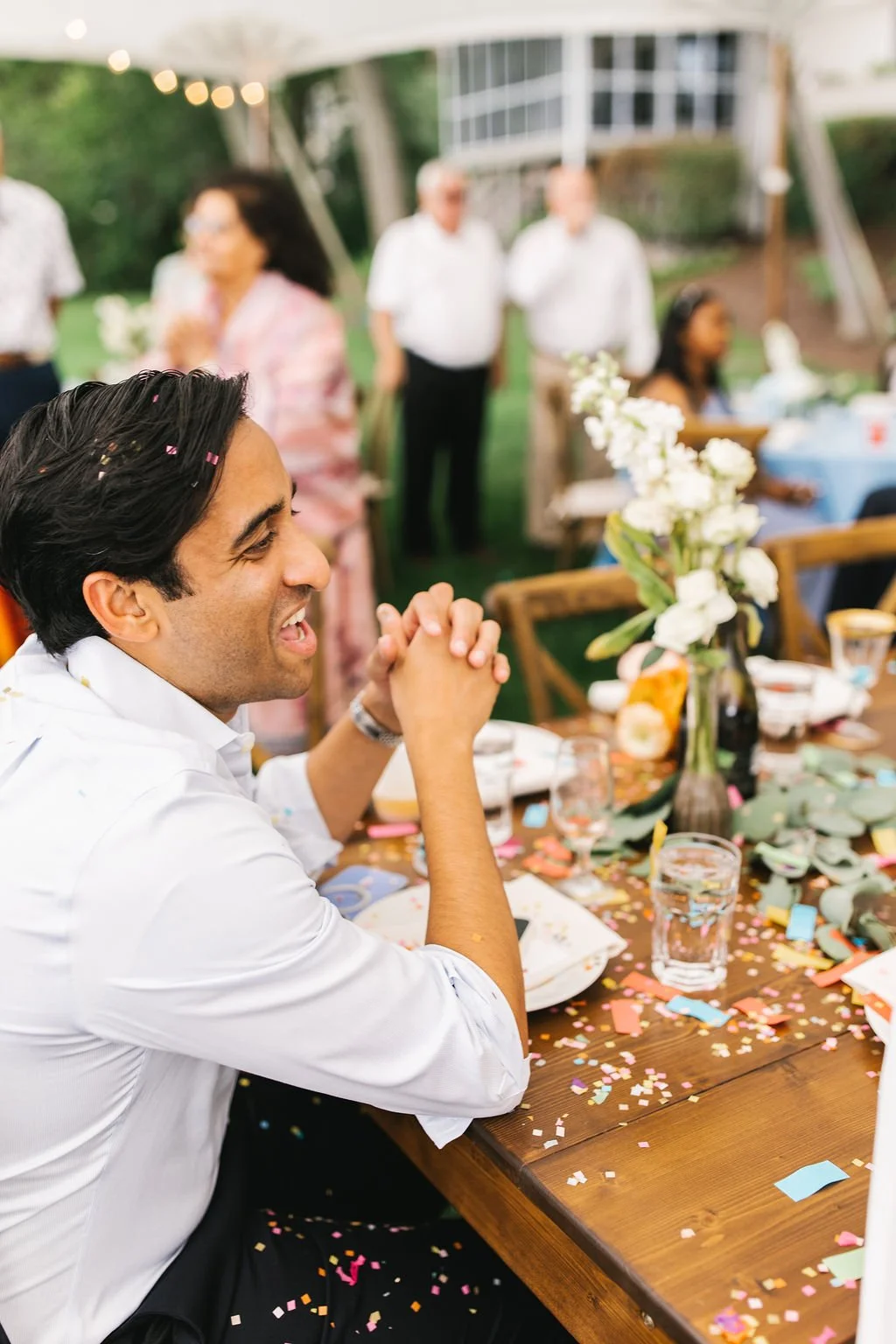 A man smiling and seated at a dining table decorated with a vase of white flowers and colorful confetti, during an outdoor celebration with people in the background.