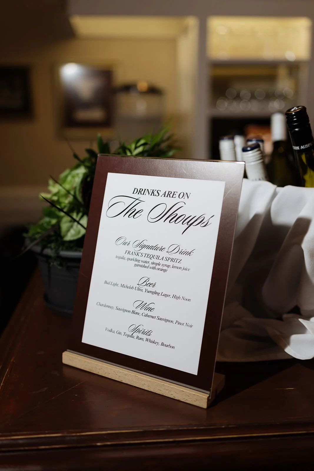 A sign listing the drink options at a wedding reception, with a bouquet of flowers nearby and bottles of alcohol in the background.