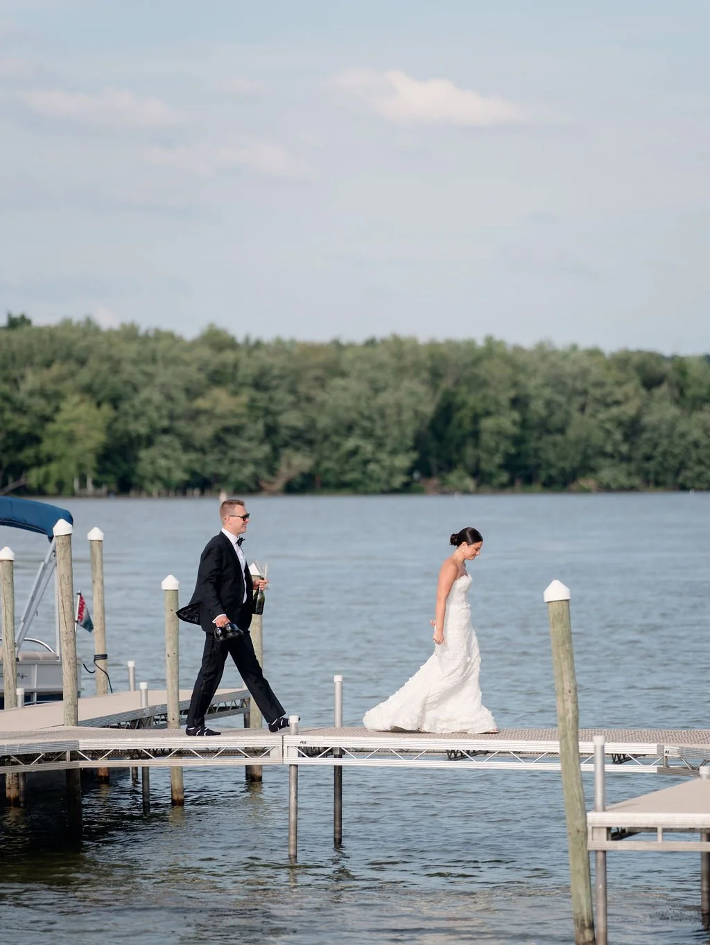A bride in a white wedding dress walking on a dock near a body of water, accompanied by a man in a black suit holding a camera, with trees and a cloudy sky in the background.