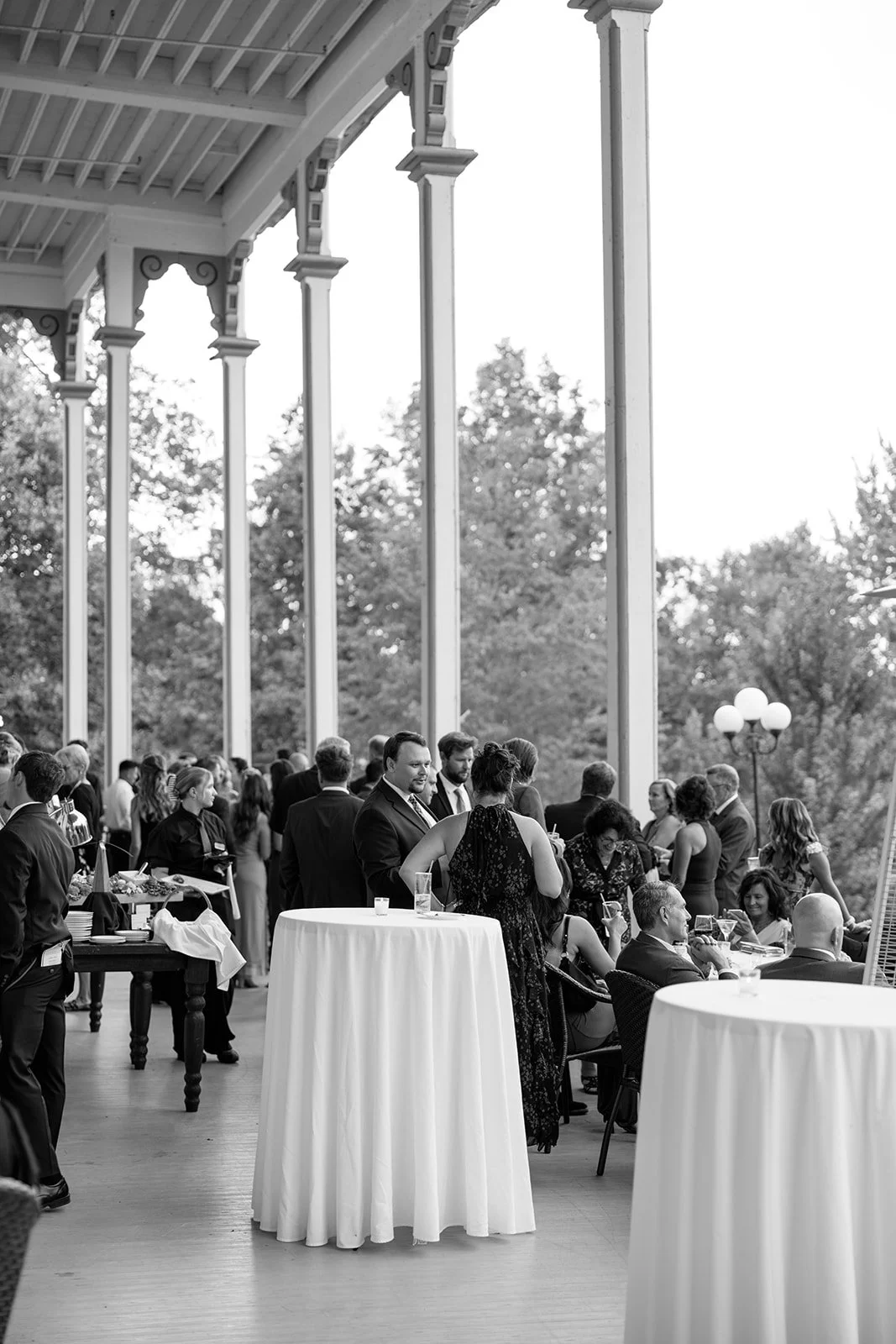 People socializing at a formal outdoor event on a veranda with tall columns and trees in the background, some standing and others seated at tables, dressed in formal attire.