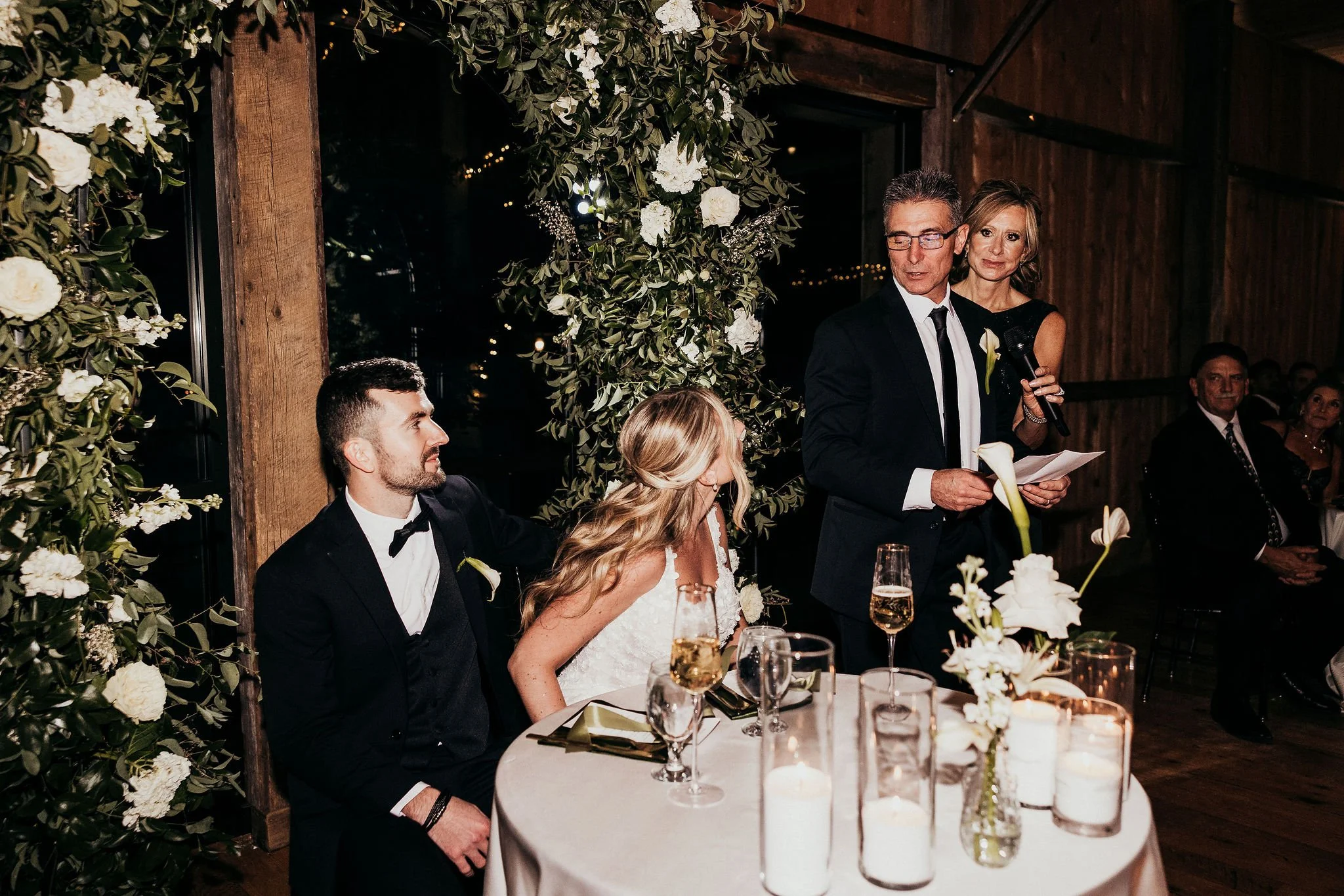 A wedding reception with a bride and groom seated at a decorated table, a man in a suit giving a speech, and guests watching. The scene features floral decorations and dim lighting.
