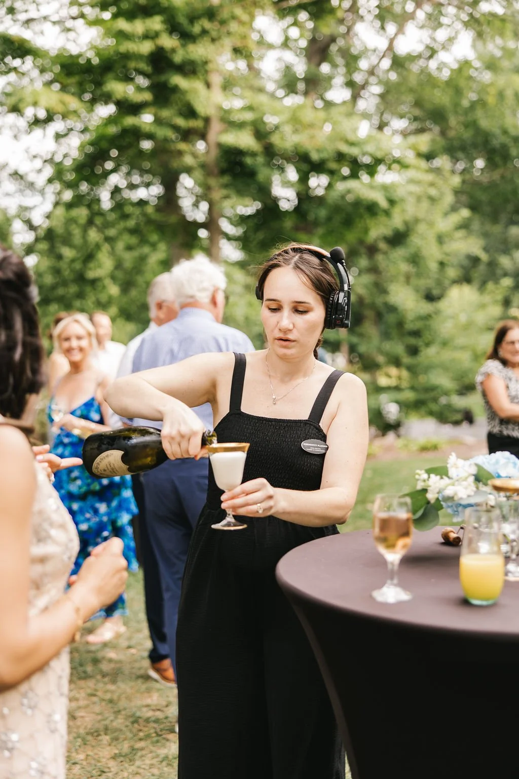 A woman with dark hair, wearing a black dress and a headset, pours champagne into a glass at an outdoor event, with other people and a table with drinks and flowers in the background.