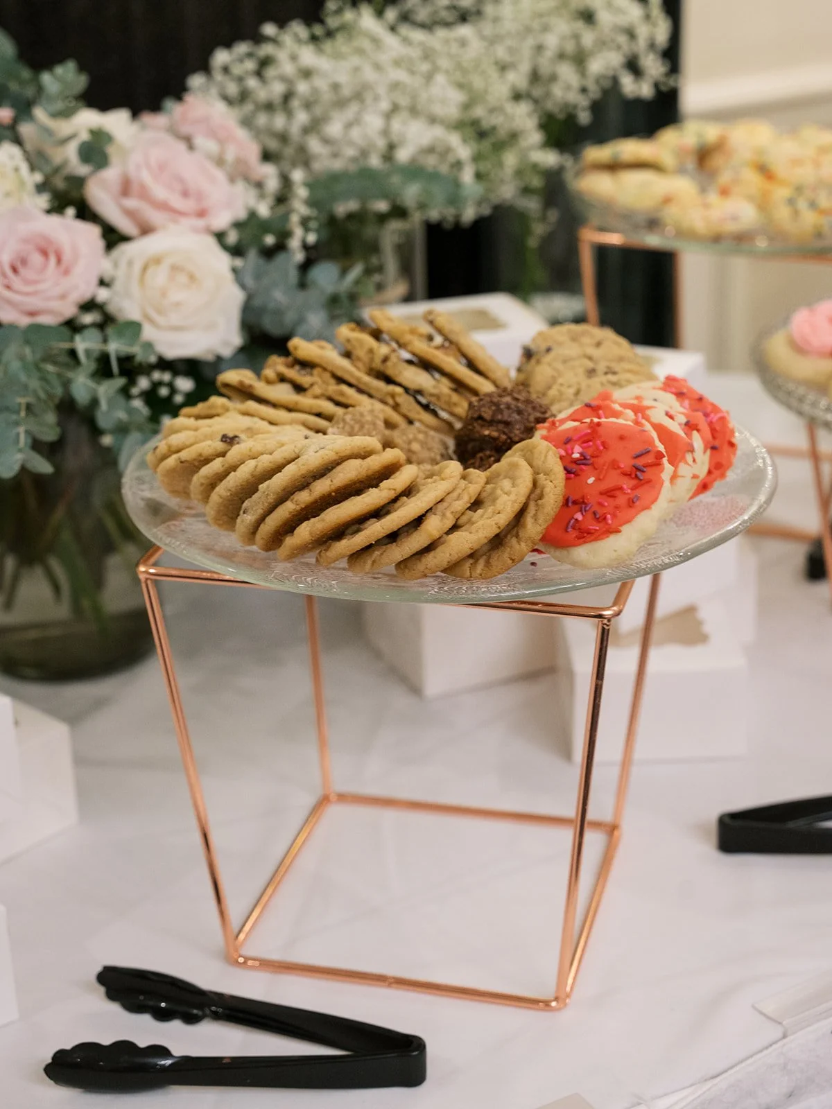 Assorted cookies and cupcakes on a glass cake stand with a rose and baby's breath floral arrangement in the background.