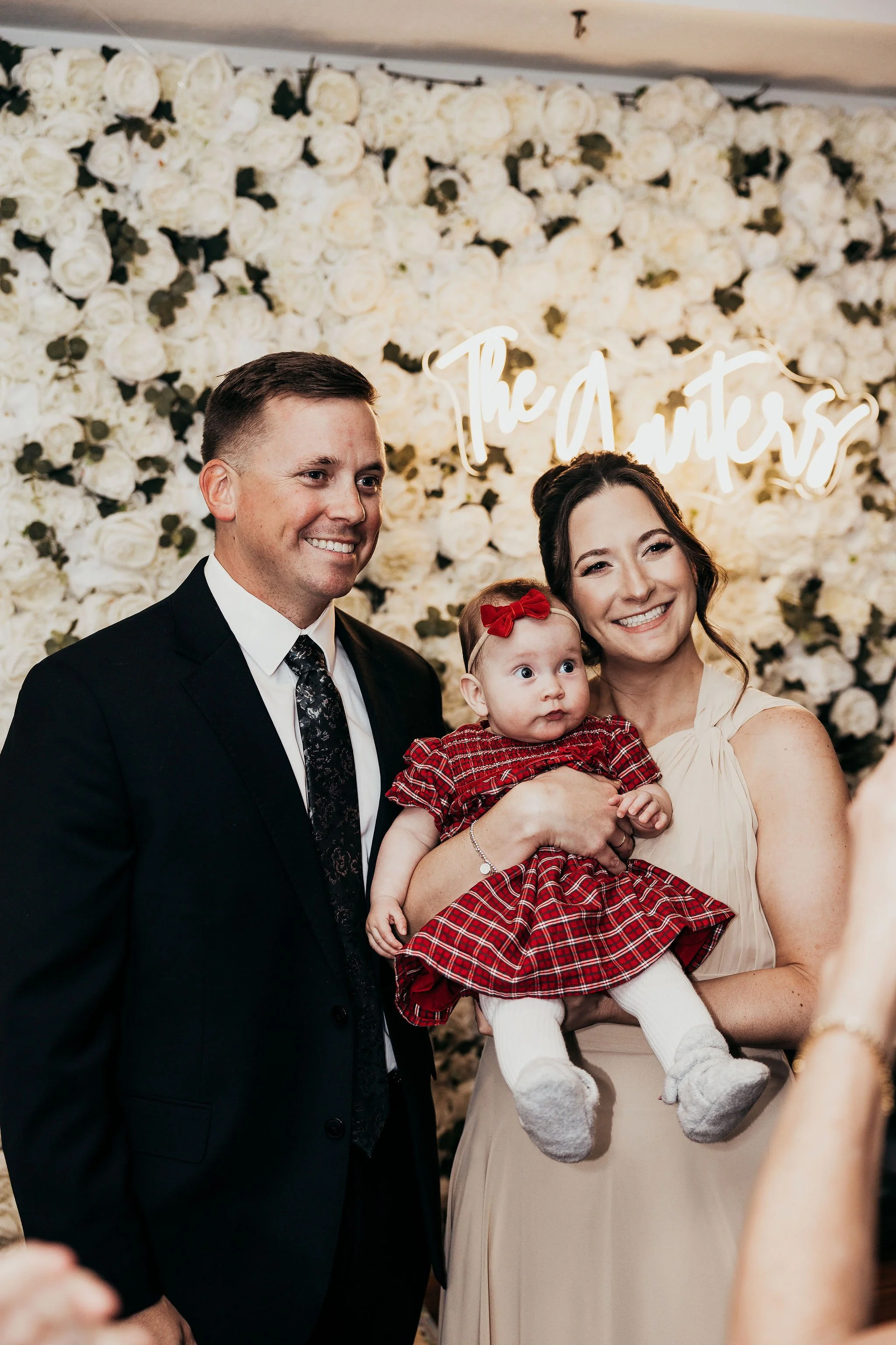 Family photo at a wedding reception with a decorated white floral backdrop and neon sign, featuring a man in a suit, a woman holding a baby girl dressed in red plaid, and smiling majority.