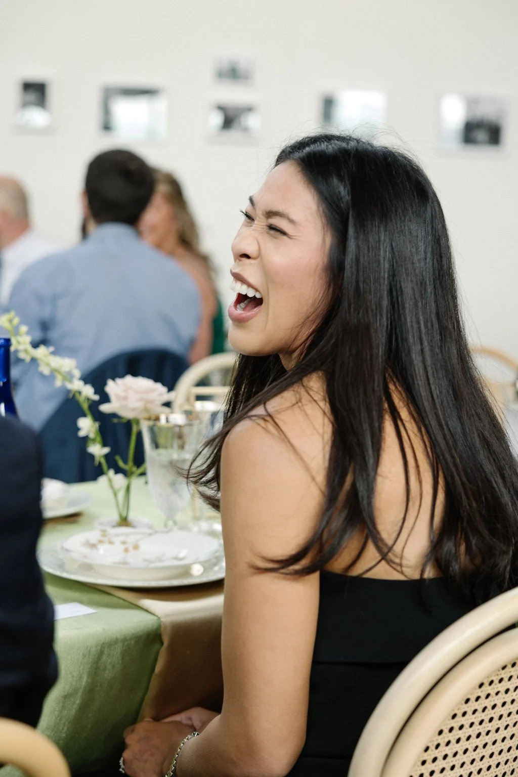 A woman with long black hair, laughing or shouting, at a celebration or gathering, with other people in the background and decorated tableware.