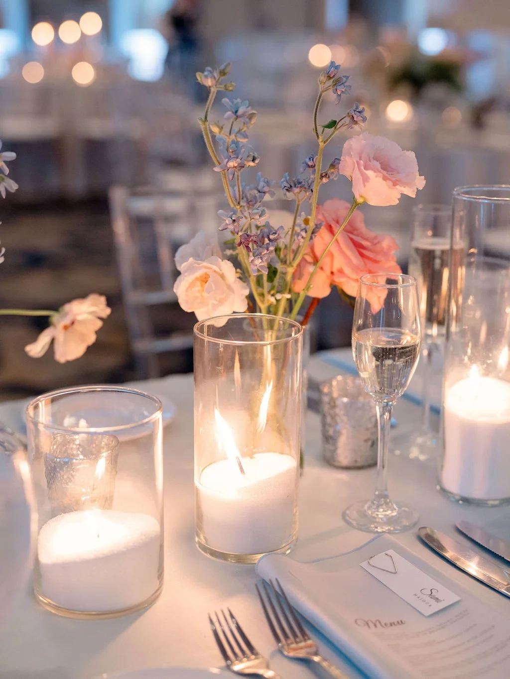Elegant table setting with lit candles, pink and purple flowers in a vase, champagne glasses, and a menu.