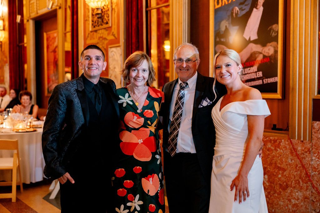 Four people posing together at a formal event with a decorated interior, including framed posters on the wall. The group includes two men and two women dressed in formal attire.