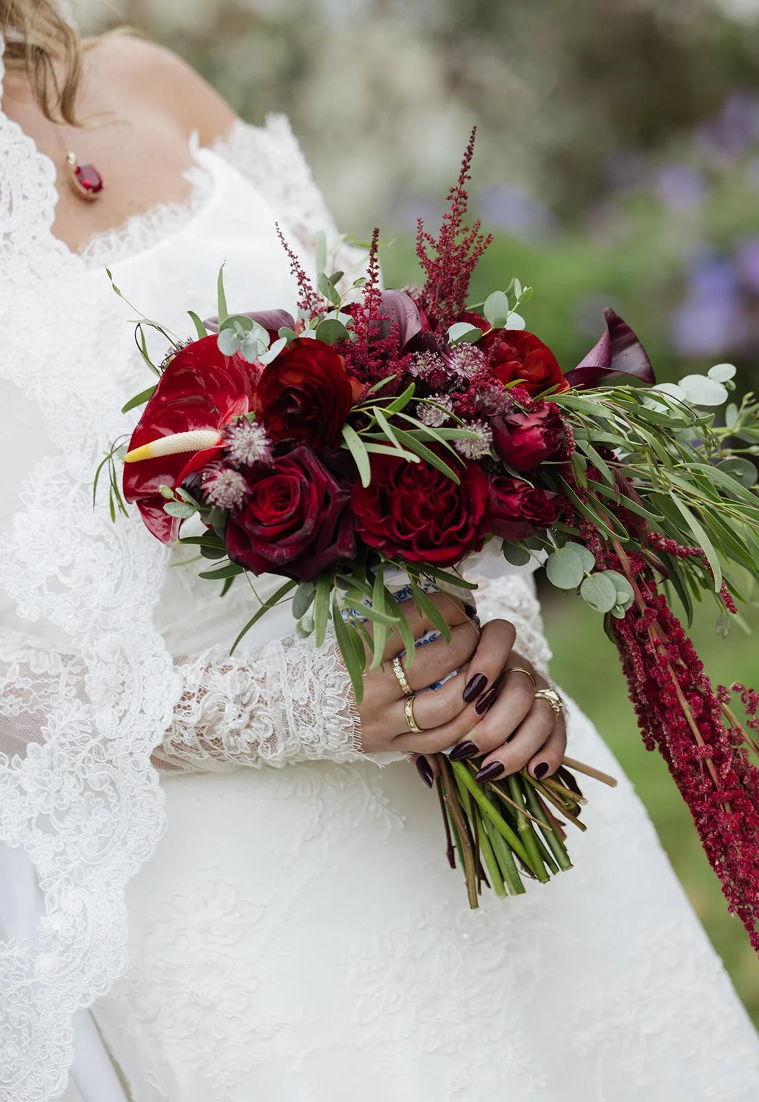A bride holding a bouquet of red roses, burgundy calla lilies, and greenery, wearing a white lace wedding dress with lace sleeves and a ruby necklace.