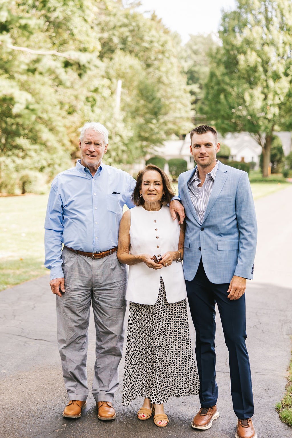 A group of three people, a man, woman, and younger man, standing outdoors on a sunny day with trees and houses in the background.