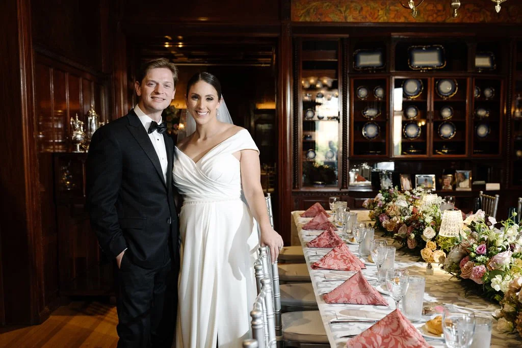 A bride and groom in wedding attire standing beside a dining table with floral arrangements and pink napkins.