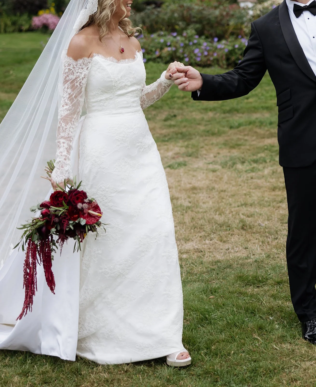 A bride and groom holding hands outdoors during their wedding ceremony, with the bride holding a bouquet of red flowers and wearing a white lace wedding dress with an off-the-shoulder neckline and lace sleeves.