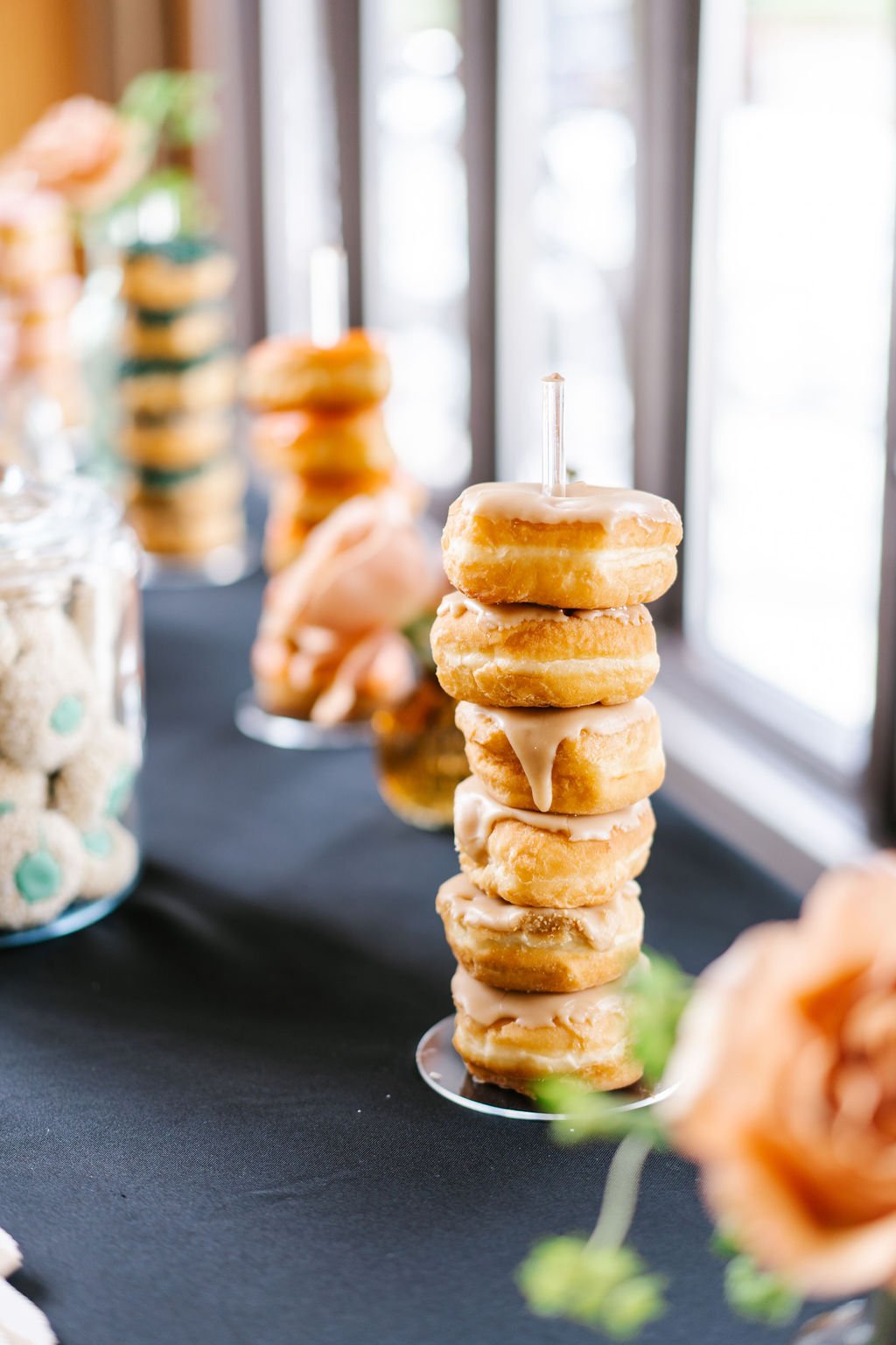 A tower of glazed donuts with a candle on top on a black table, with jars of macarons and other sweets blurred in the background.