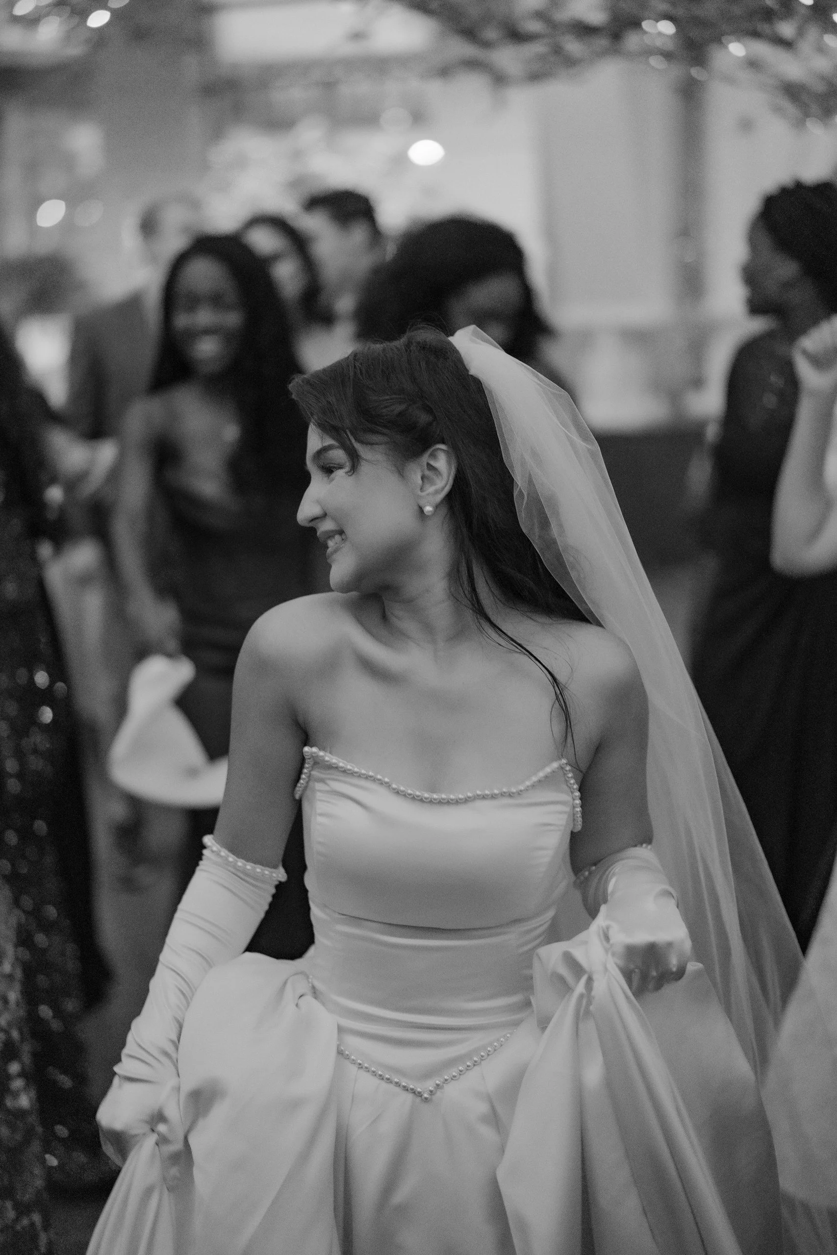 A Black and White Photo of a bride with a smile, wearing a wedding gown and gloves, holding her dress with one hand, and looking downwards. There are guests in the background, some smiling, at what appears to be a wedding reception.