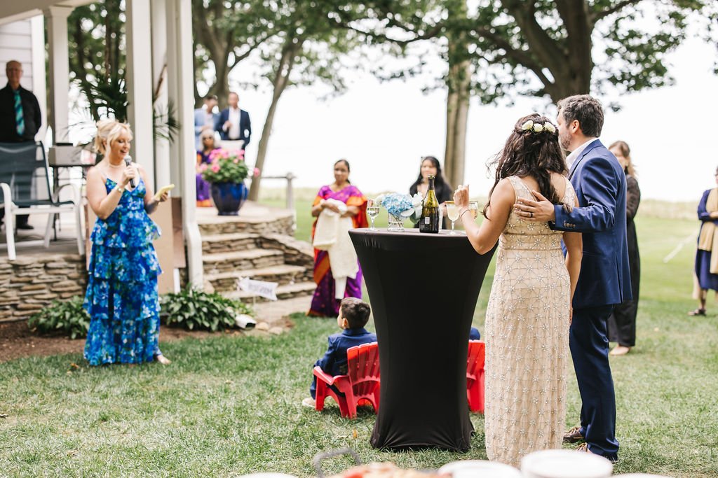 A wedding reception outdoors with a bride and groom dancing, a woman in a blue floral dress giving a speech, and guests in colorful attire watching.