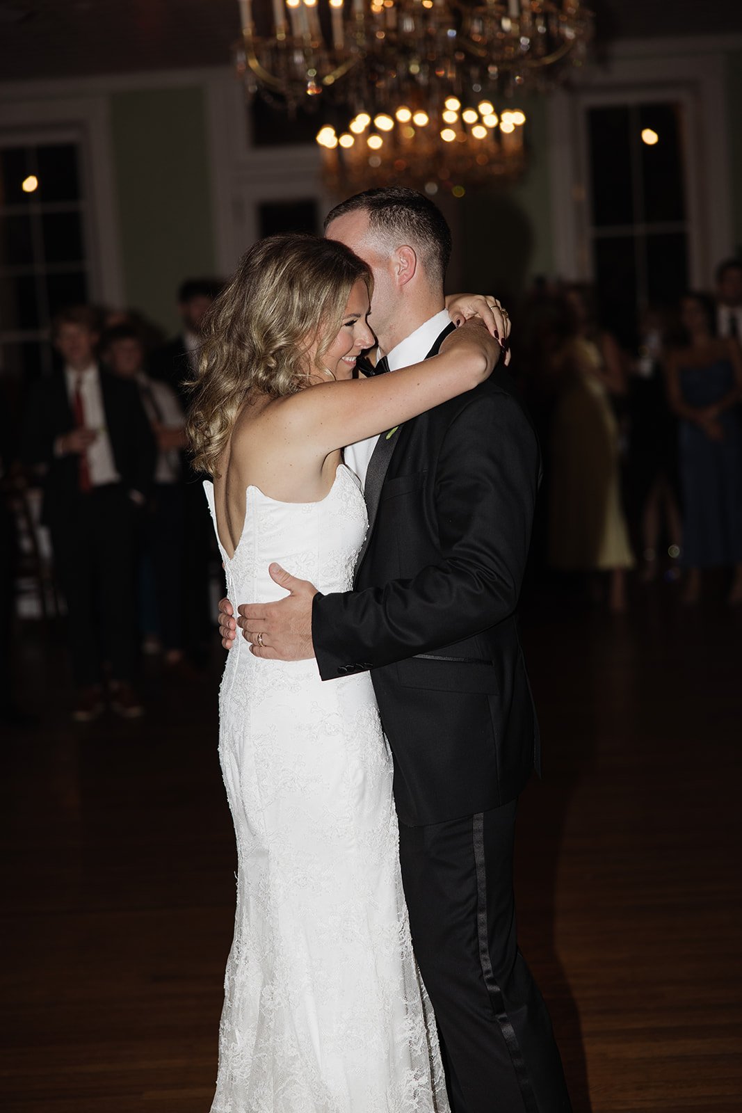 A bride and groom are dancing closely at their wedding reception, surrounded by guests in the background.