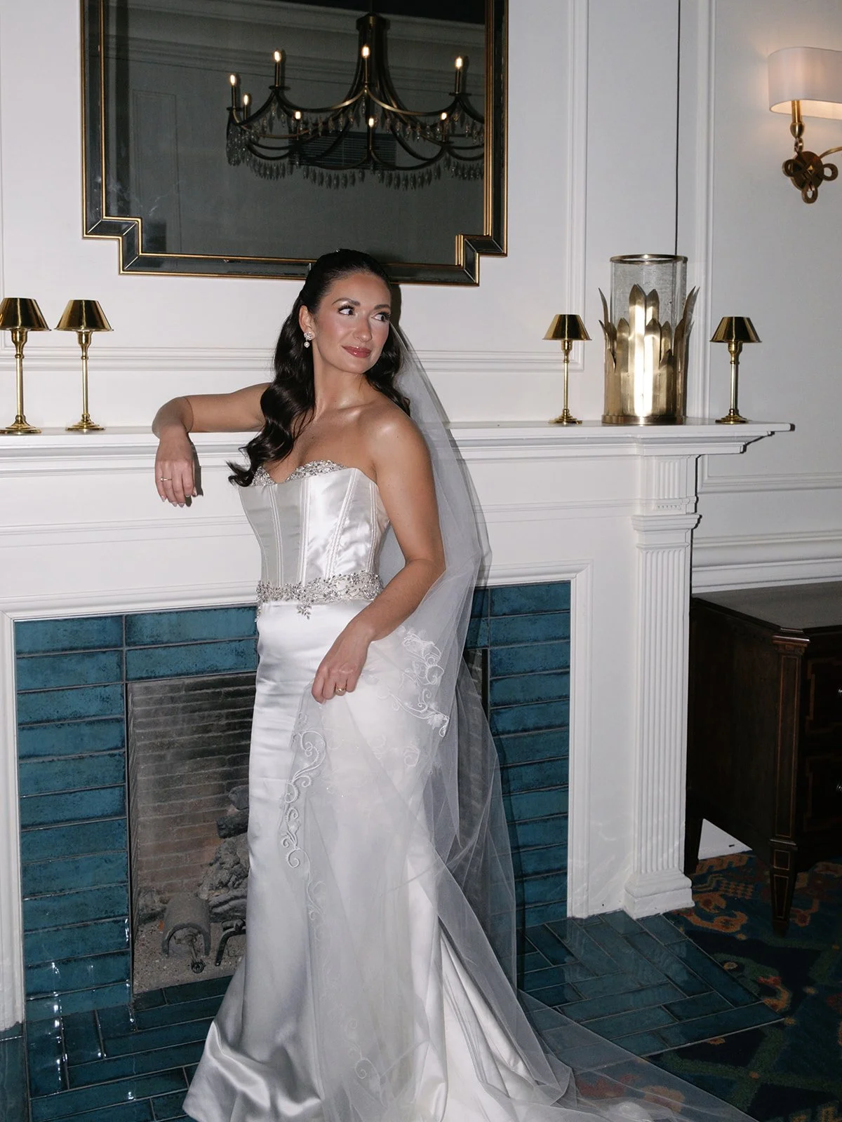 A woman in a white wedding gown standing next to a fireplace with a mirror and gold accents in the background.