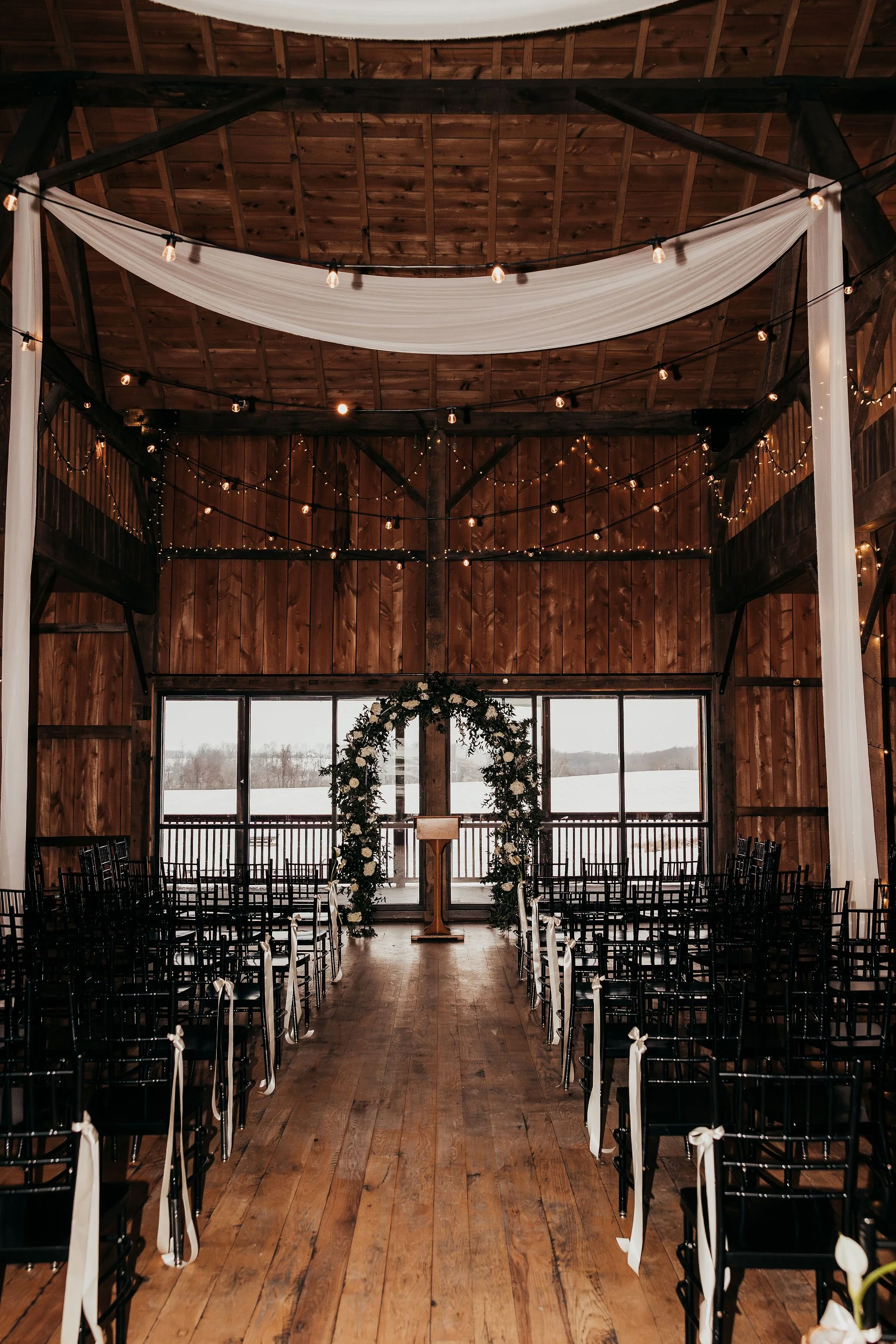 Indoor wedding ceremony setup with chairs decorated with white ribbons, an arch with white flowers, and string lights hanging from a wooden ceiling inside a rustic venue.