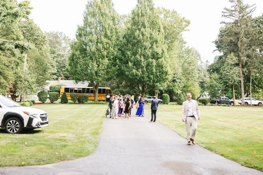 People gathered outside on a paved pathway surrounded by green grass and trees, with cars parked on the side and a school bus in the background, during daytime.