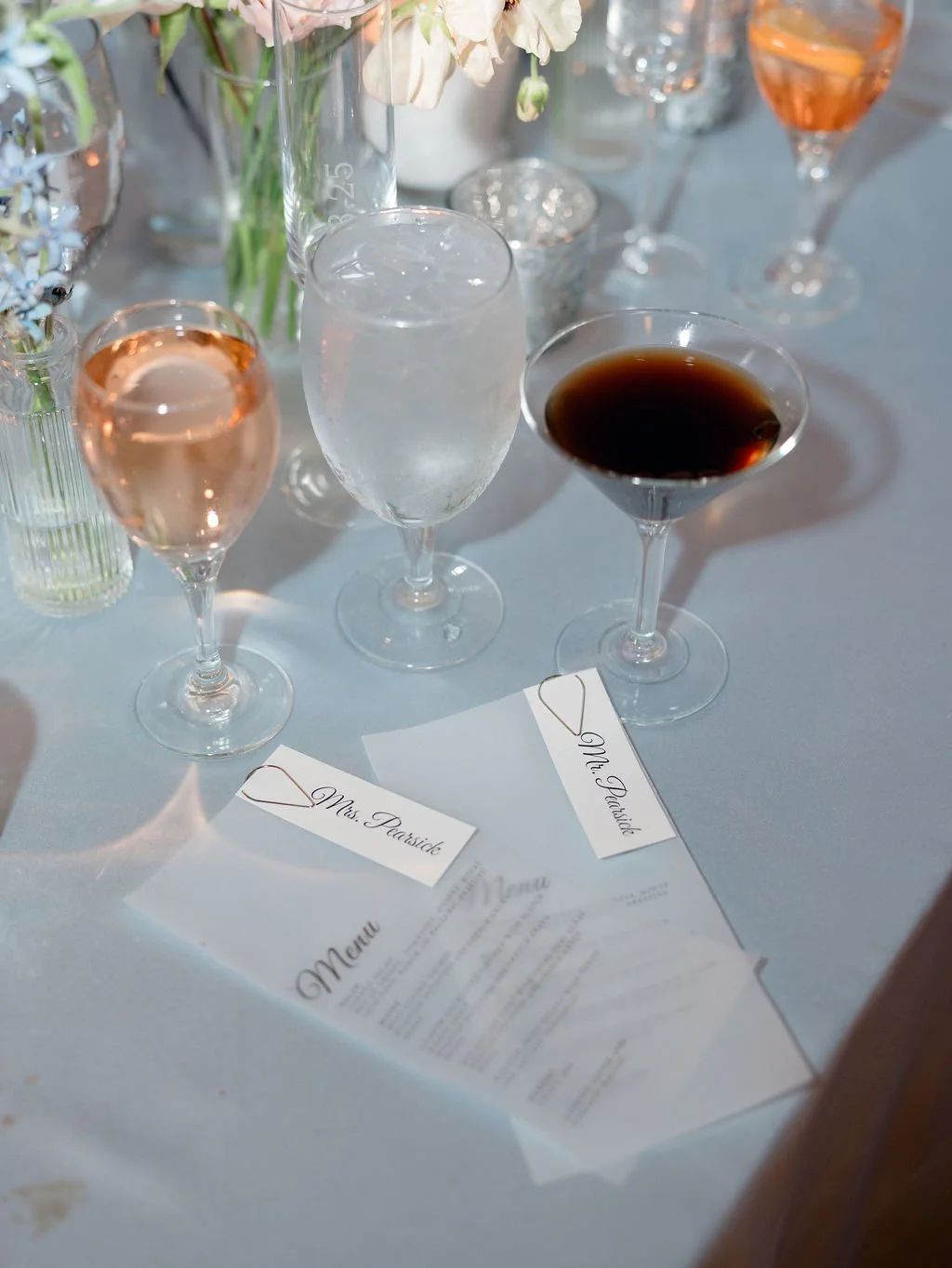 Table with glasses of water, and various alcoholic drinks such as rosé wine, sparkling wine, and red wine, along with a menu and name cards for "Mrs. Purdie" and "Mr. Purdie". There are flower vases in the background.
