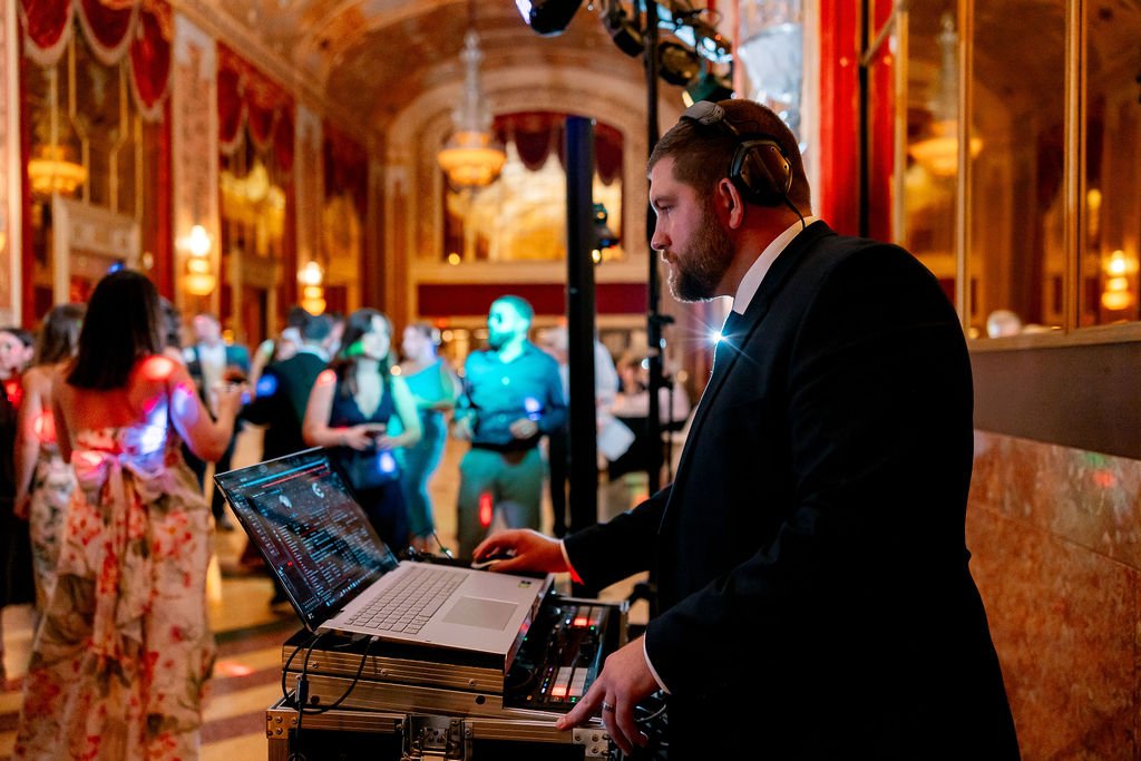 A DJ in a black suit is playing music on a laptop and DJ controller in a grand ballroom with ornate gold decorations and chandeliers, while a crowd of people dances in the background.