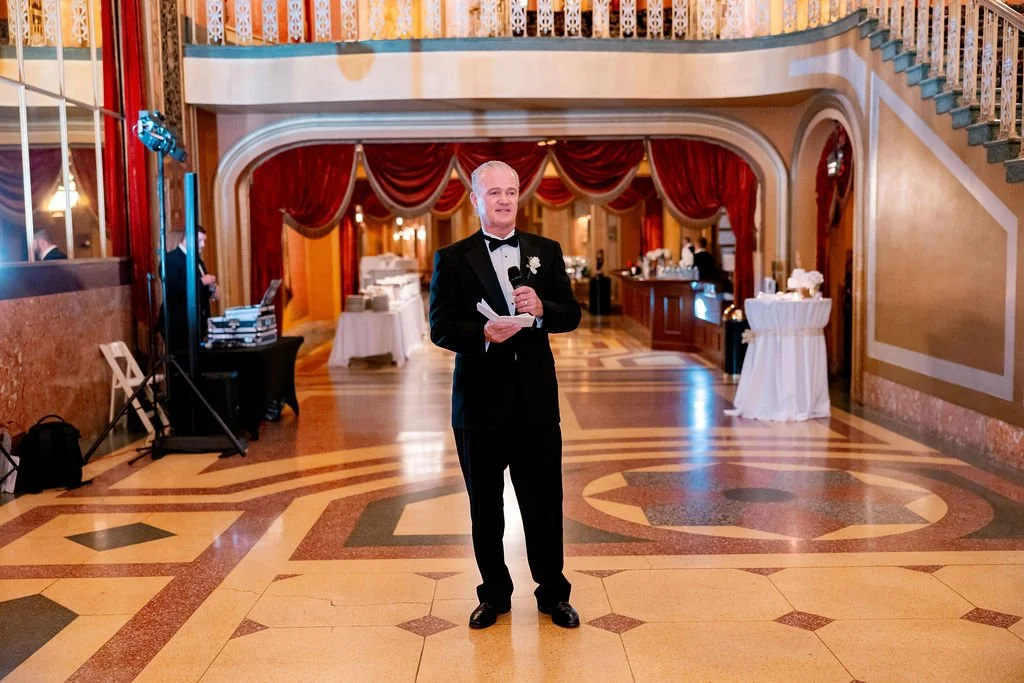 A man in a tuxedo speaking into a microphone at a wedding reception in an elegant hall with hardwood floors, red curtains, and a staircase.