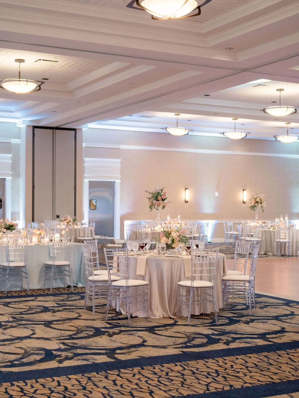 Elegant banquet hall decorated for a wedding reception with round tables, floral centerpieces, and clear chairs, featuring soft lighting and a patterned carpet.