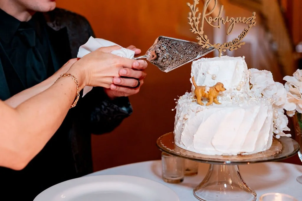 Person cutting a white wedding cake with a silver cake knife, decorated with a small dog figurine and a gold topper that reads "Rearguez" with a date, on a gold-colored cake stand