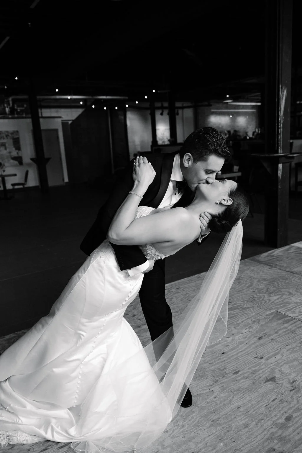 A newlywed couple sharing a romantic kiss, with the groom dipping the bride, in a black-and-white photograph. The bride is in a wedding gown with a veil, and the groom is in a tuxedo.