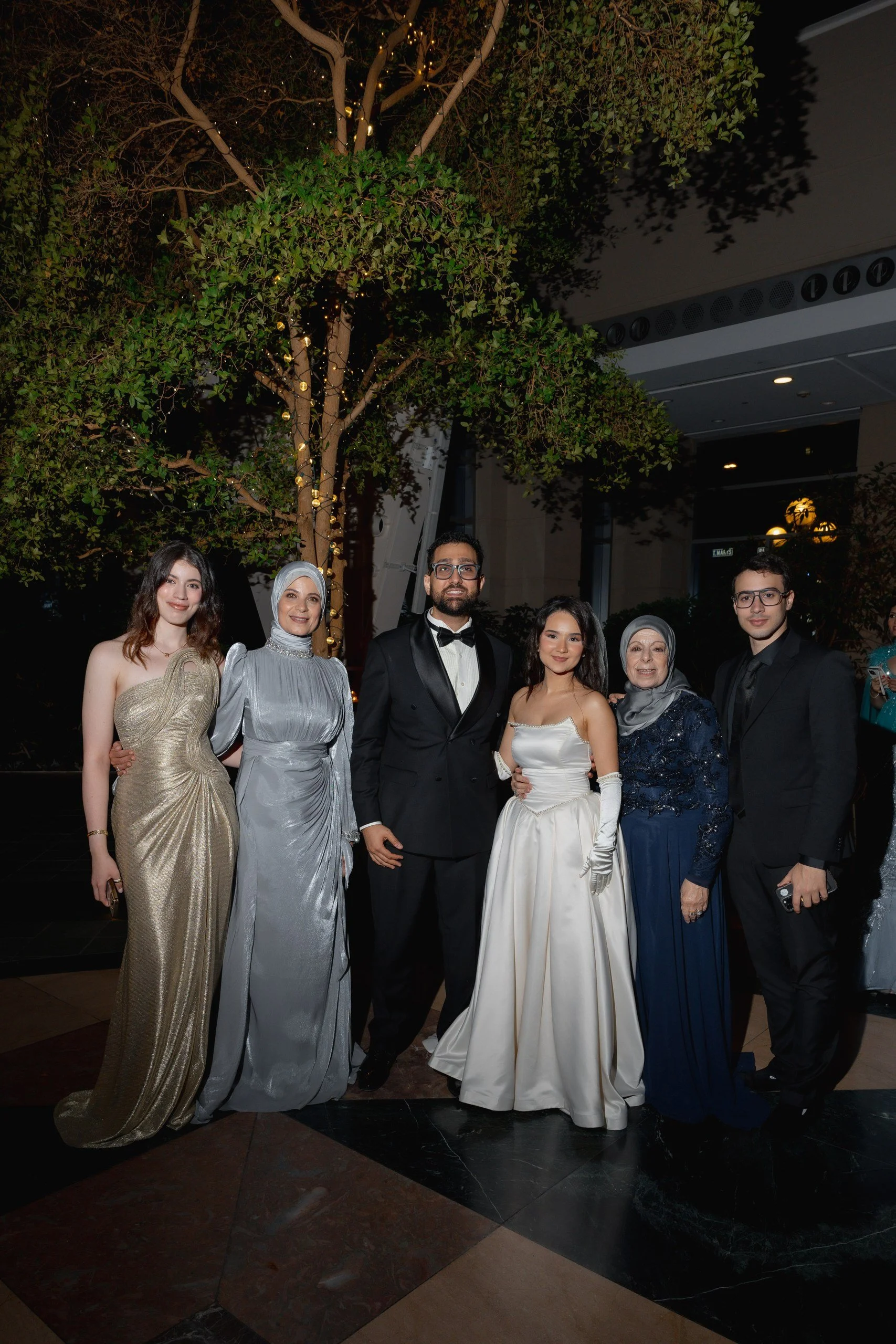 Group of seven elegantly dressed people posing at a formal event under a decorated tree with string lights, in an indoor setting.