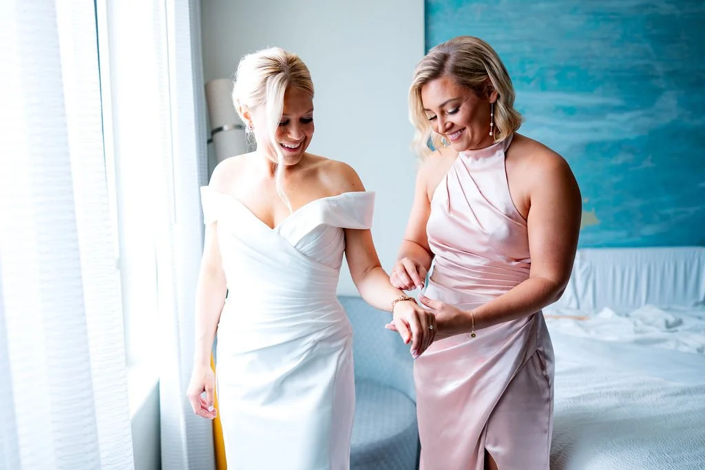 Two women, one in a white wedding dress and the other in a pink satin dress, smiling and holding hands in a bright room with a bed and blue wall in the background.