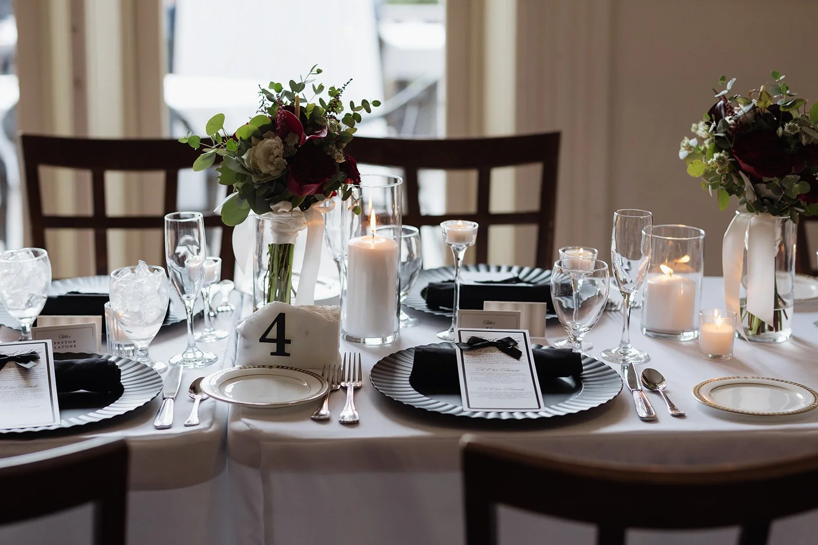 Elegant wedding table set with black and white plates, silverware, candles, floral centerpieces, and table number 4 in a well-lit room.