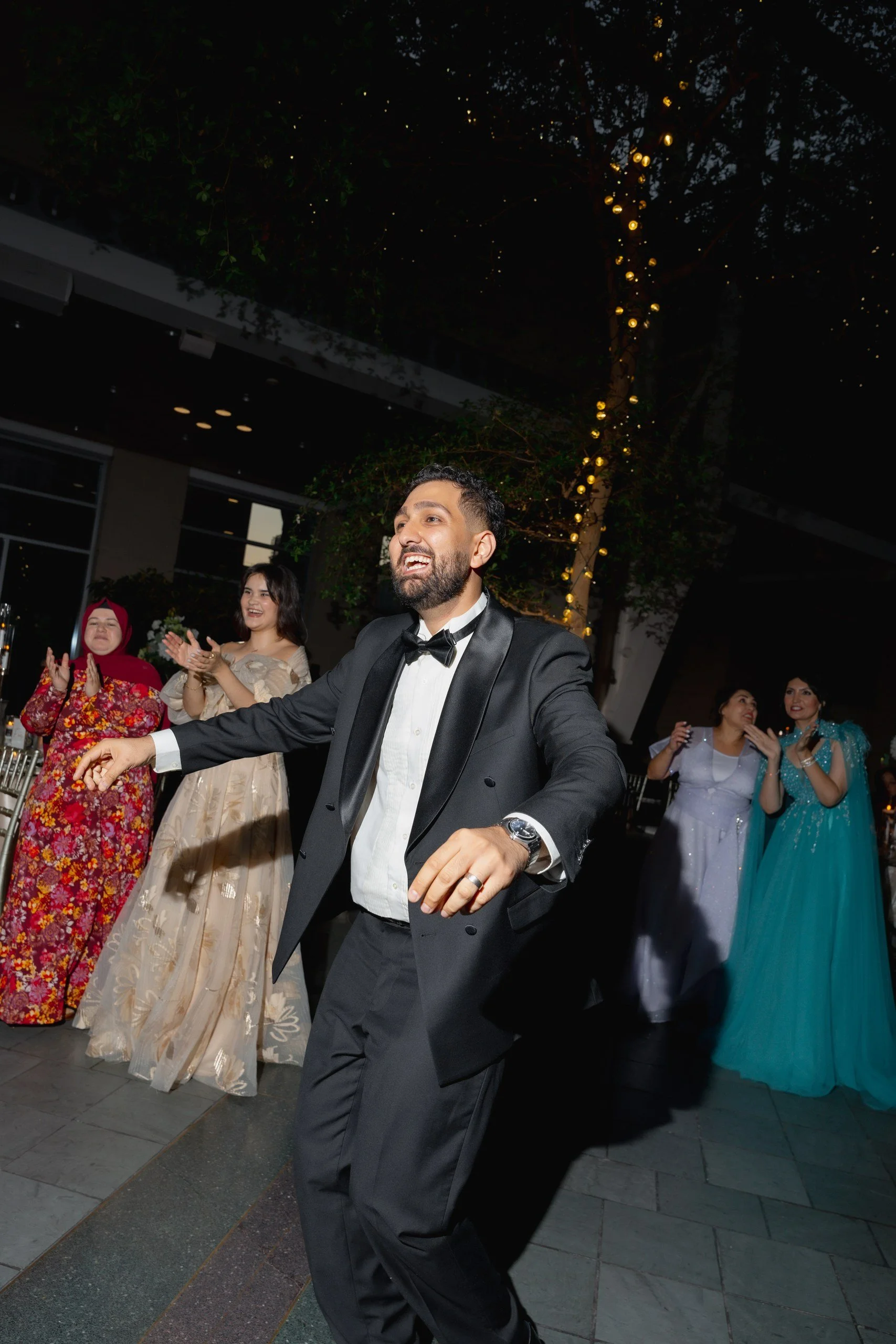 People dancing and celebrating at a formal event outdoors at night, with one man in a tuxedo in the foreground and women in colorful dresses in the background.