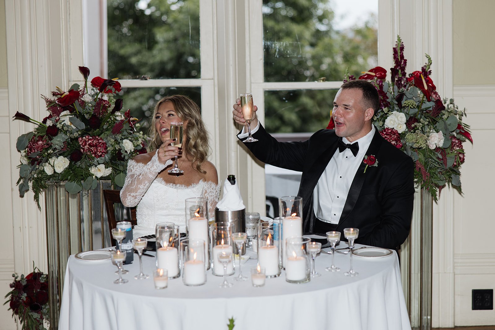 A bride and groom celebrating at a wedding reception, raising champagne glasses in a toast, seated at a table decorated with candles and floral arrangements, in front of a window with greenery outside.
