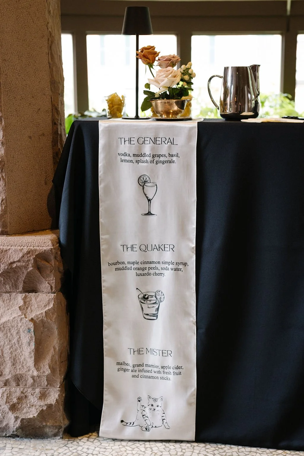 A dinner menu banner hanging on a dark tablecloth with a vase of pink and white roses, a small yellow flower, a black lamp, and a shiny metal pitcher on top. The menu lists three drink options: 'The General', 'The Quaker', and 'The Mister' with their