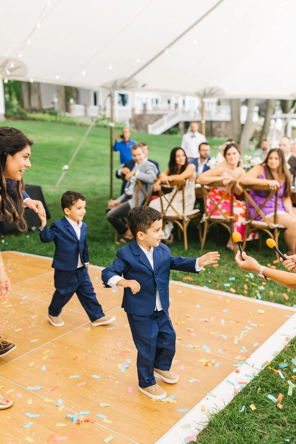Children in formal attire playing a game under a tent while being watched by adults at an outdoor event.