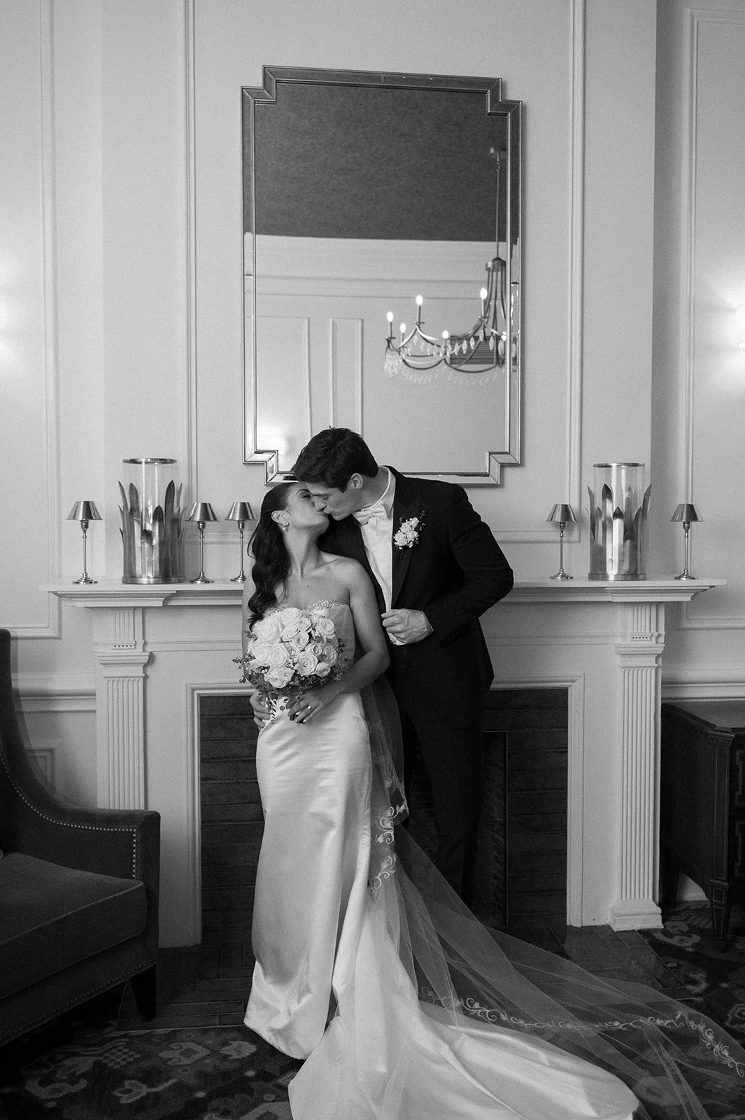 A black and white photo of a bride and groom sharing a kiss in front of a fireplace mantel with candles and vases, with a large mirror and chandelier reflecting on the wall behind them.