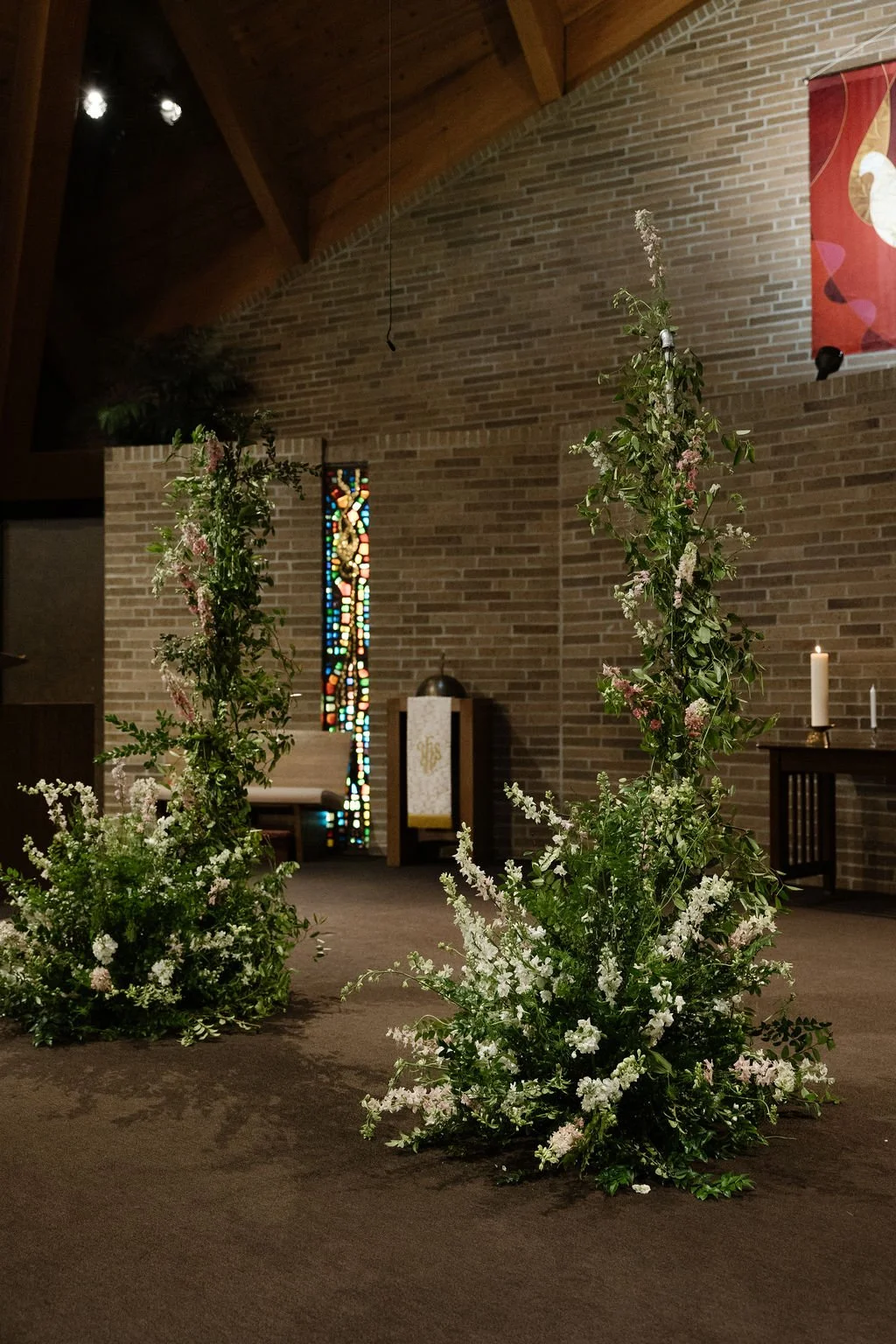 Floral arrangement with green leaves and white flowers on the floor inside a church with brick walls, stained glass window, and candles on an altar table.