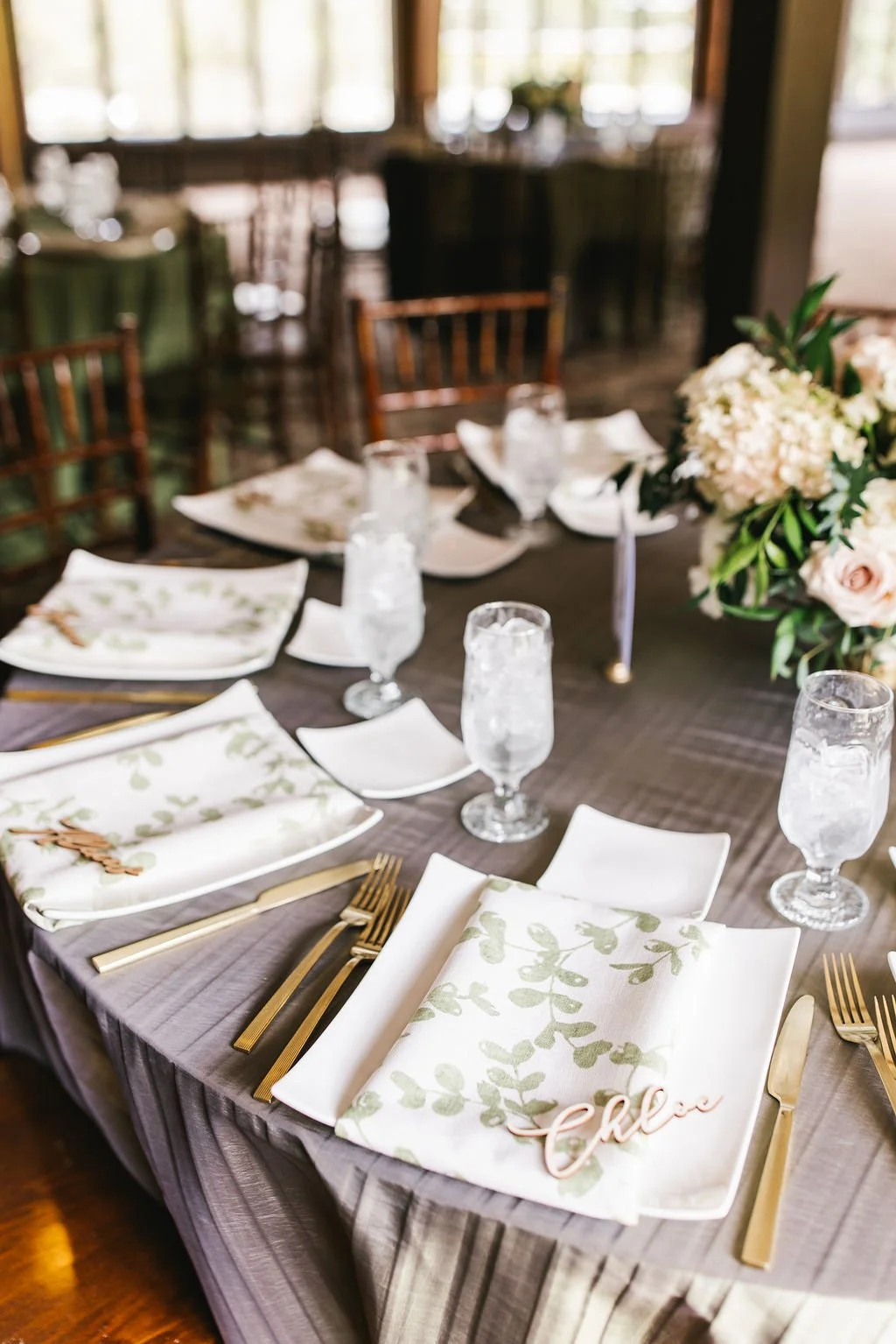 Elegant table setting with white plates, gold utensils, floral napkins, and glassware, decorated with a large bouquet of pink and white flowers at a wedding or special event.