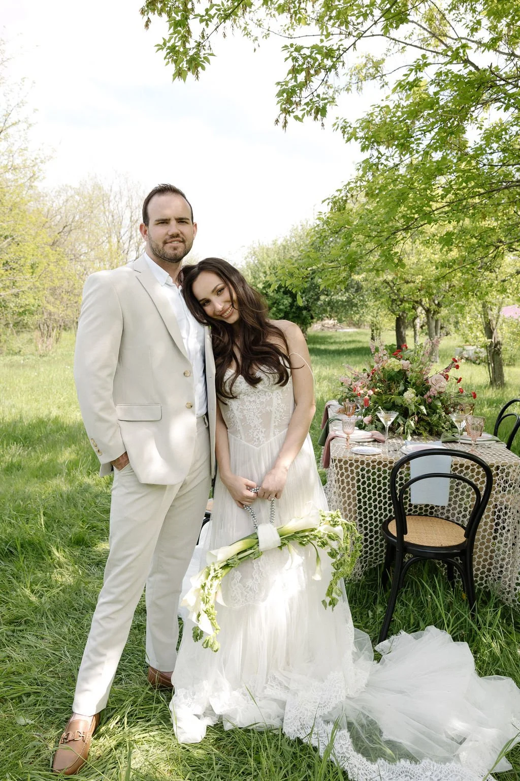 A bride and groom standing outdoors with a decorated table behind them, green trees, and grass during a wedding celebration.