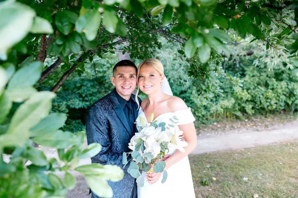 A bride and groom smiling together outdoors, surrounded by green foliage, with the bride holding a bouquet of white flowers and greenery.