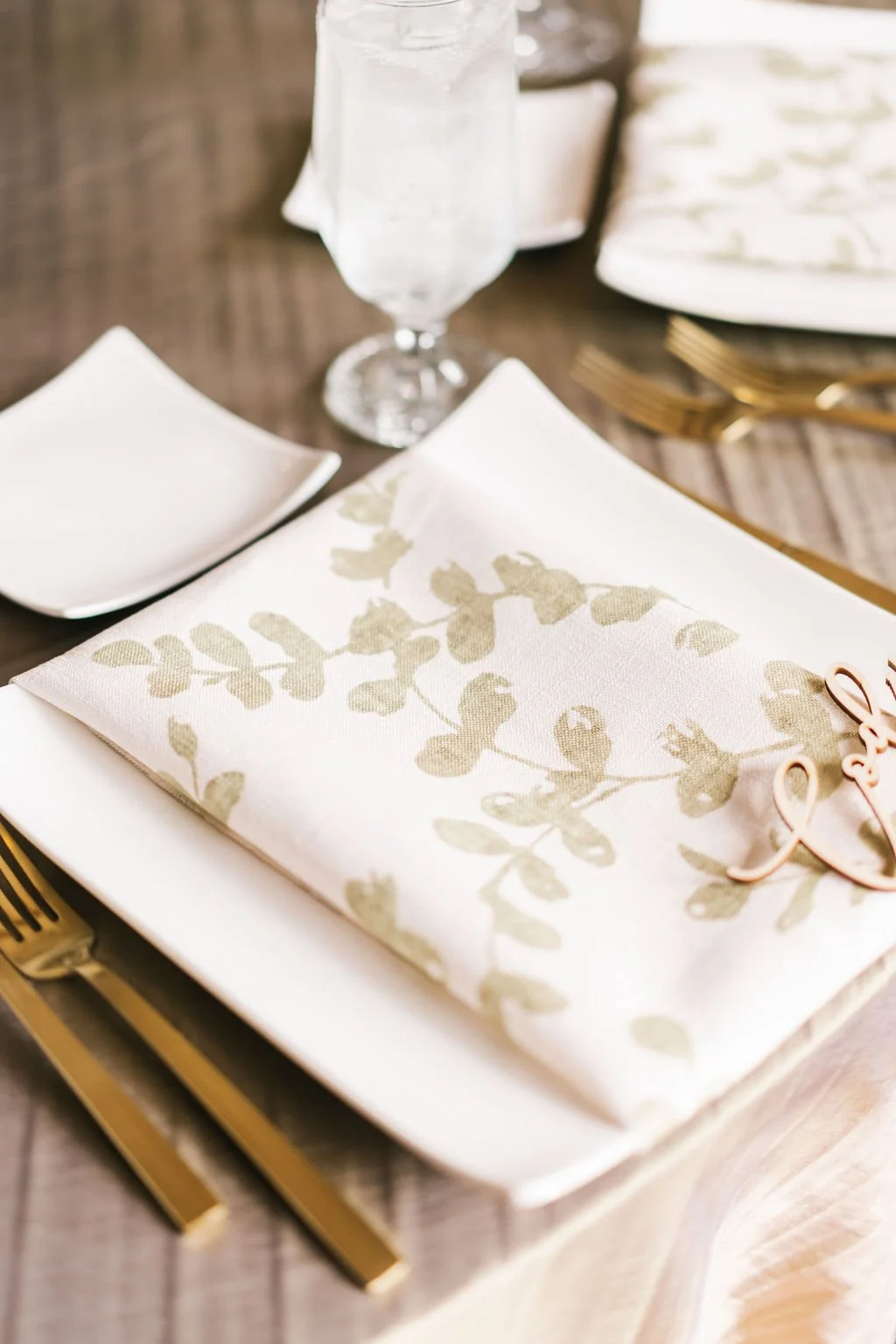 Elegant table setting with a white plate, gold flatware, a floral napkin, and a glass of water on a wooden table.