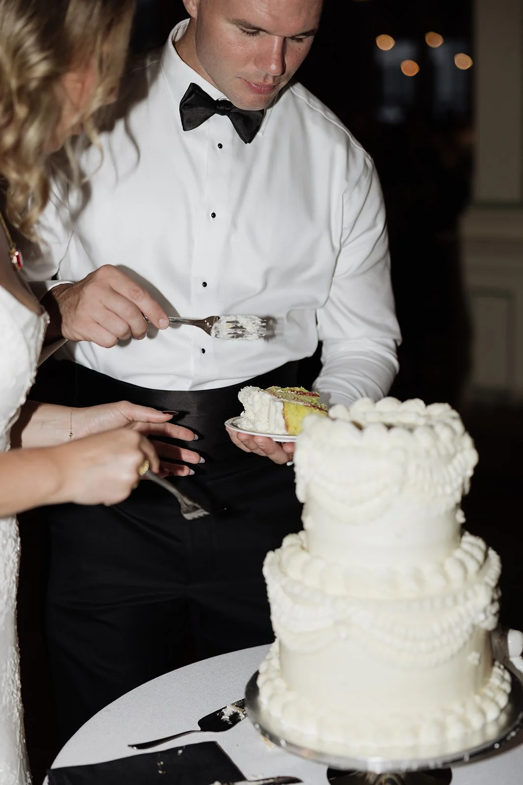 A man dressed in formal attire, including a white shirt and black bow tie, is cutting and serving a slice of wedding cake at an event.