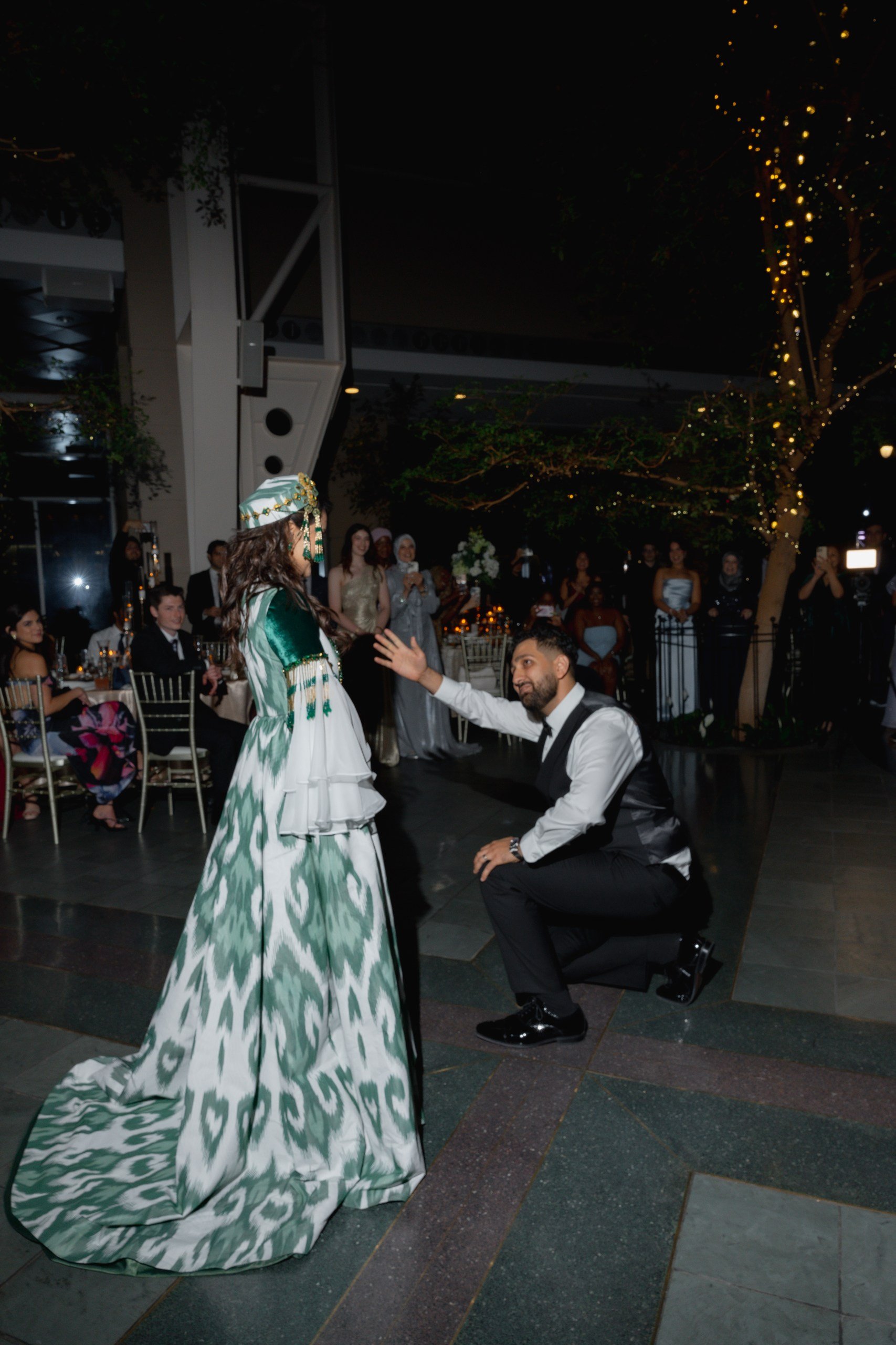 A couple is engaged in a marriage proposal at a nighttime outdoor event, with the man kneeling and holding the woman's hand. The woman is dressed in a green and white patterned gown and wearing a matching headpiece, while the man is dressed in a whit