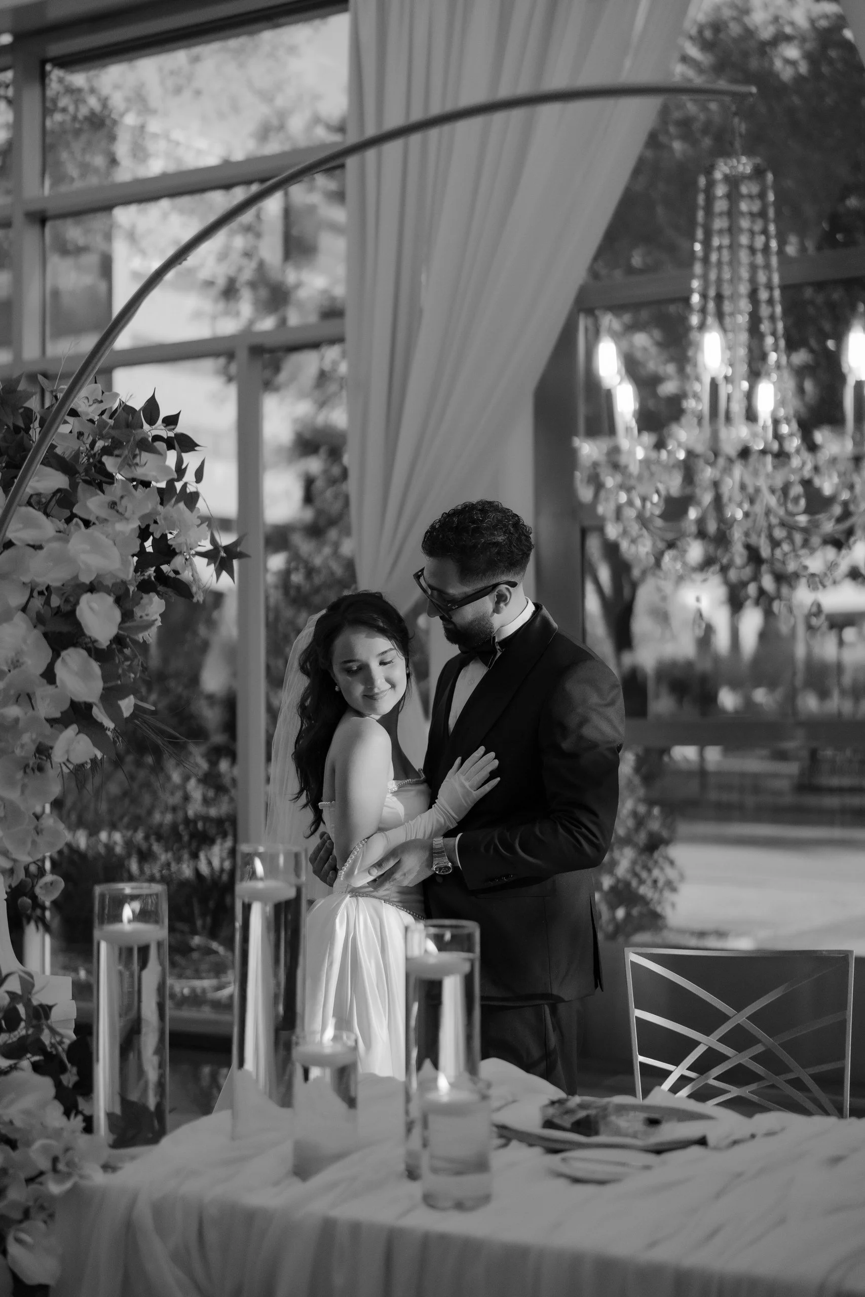 A black and white photo of a bride and groom dancing together at their wedding reception. The bride is wearing a white dress and veil, and the groom is in a tuxedo. They are holding each other, with the bride smiling and the groom looking at her. The