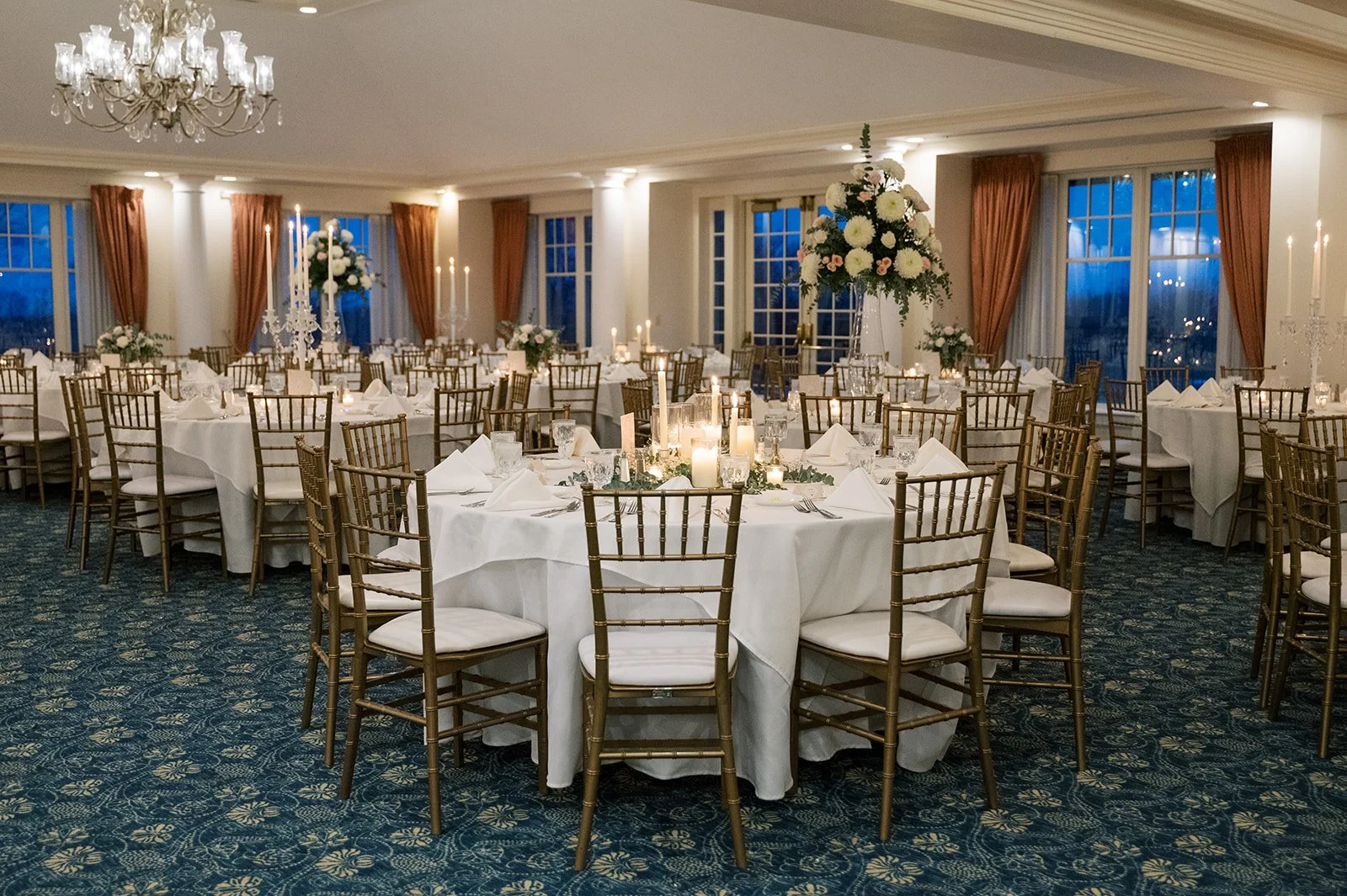 Elegant banquet hall decorated for a formal event with white tablecloths, gold chairs, tall floral centerpieces, and tall candle arrangements. Large windows with orange curtains, chandelier, and blue evening sky visible outside.